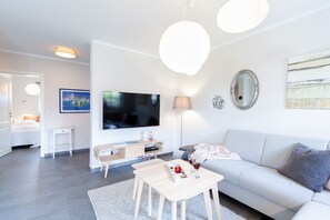 Ein modernes Wohnzimmer mit einem Flachbildfernseher, einer Couch, einem Couchtisch und einem Essbereich mit einer Lampe und einem kleinen Tisch.