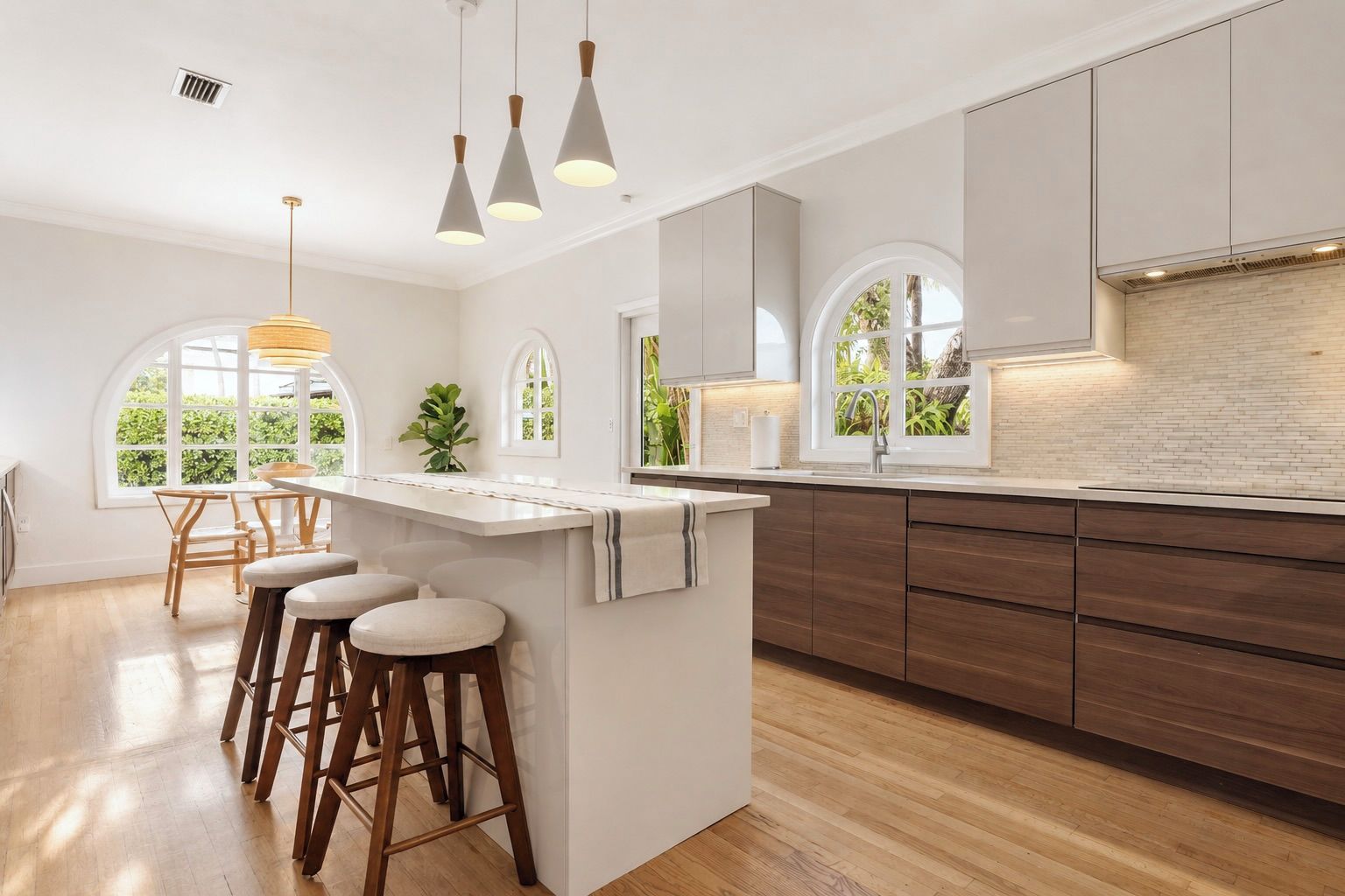 A modern kitchen with a central island, bar stools, pendant lighting, and a view of greenery through large windows.