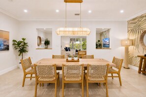 A dining area with a wooden table and woven chairs, a hanging light fixture, and a view of greenery outside.