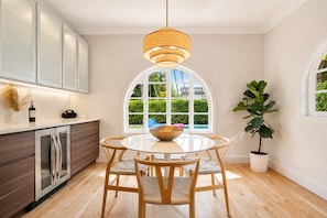 A modern kitchen with a round dining table, wooden chairs, a hanging light fixture, and a view of a swimming pool through a large arched window. - Brickell Oasis | Heated Pool, Fire Pit + Mini Golf (Miami)
