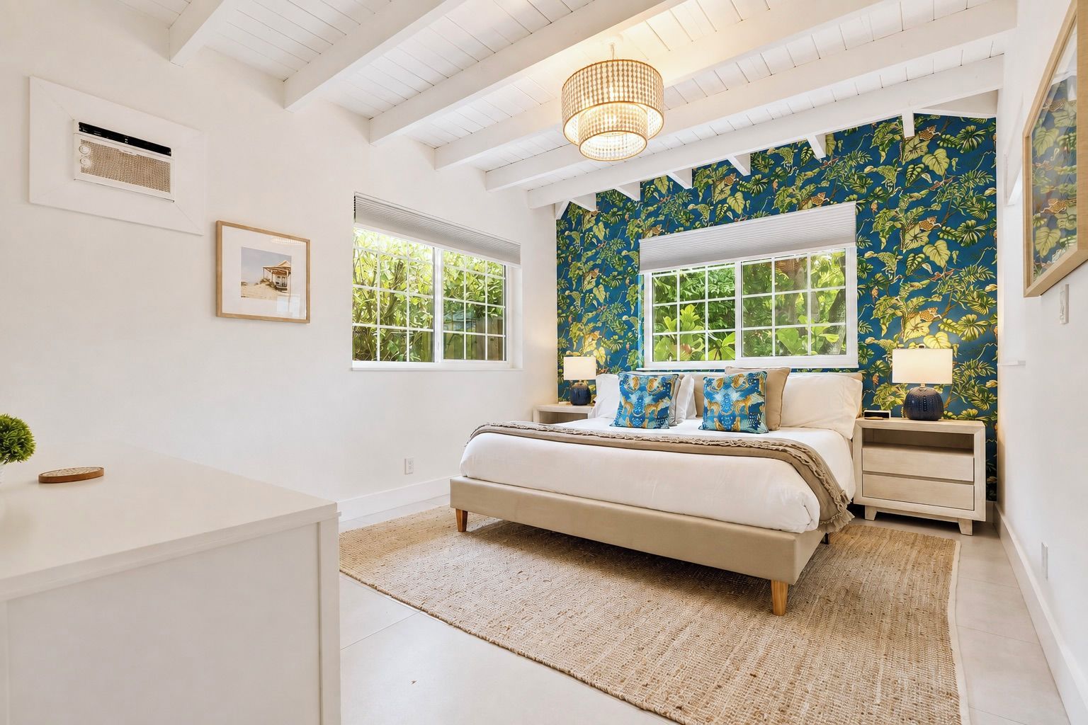 A bedroom with a bed, a nightstand, a lamp, a window, and a wall with a tropical leaf pattern.