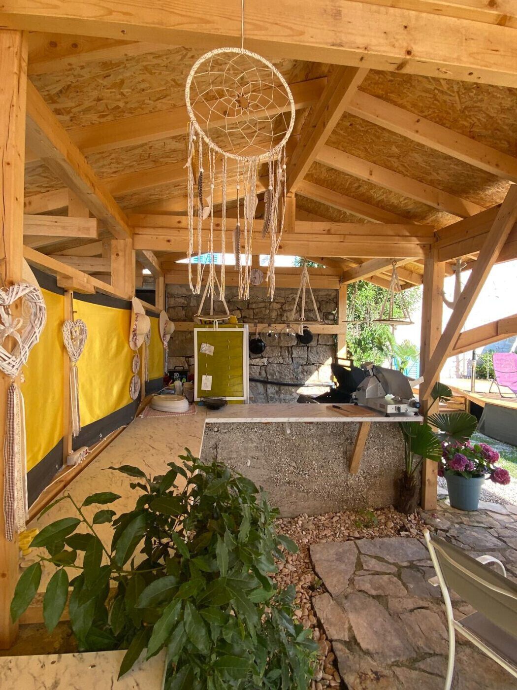A rustic wooden structure with a stone wall, a yellow wall with decorative items, and a hanging dreamcatcher.