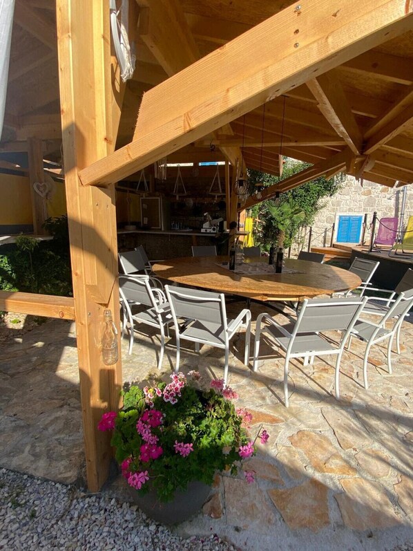 An outdoor dining area with a wooden pergola, stone flooring, and metal chairs.