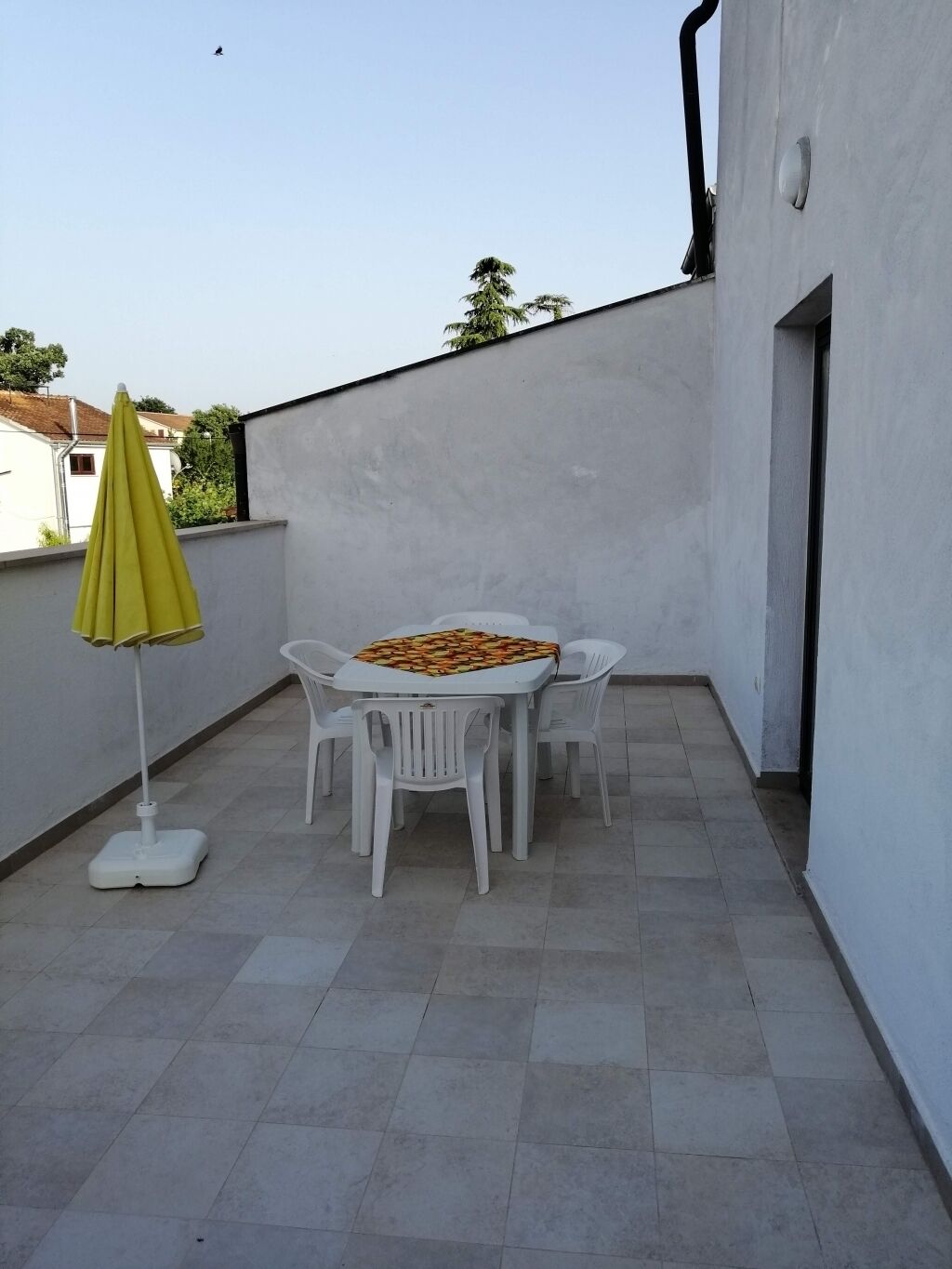 Een klein terras buiten met een witte tafel en stoelen, een gele parasol en een witte vloerlamp.