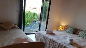 A hotel room with two beds, a chair, a small table with a lamp, and a view of the outside through a glass door.