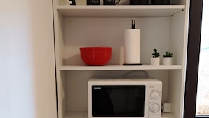 Eine Küche mit weißen Regalen, einer roten Schüssel, einer weißen Mikrowelle, einem Edelstahl-Wasserkocher und zwei kleinen Topfpflanzen.