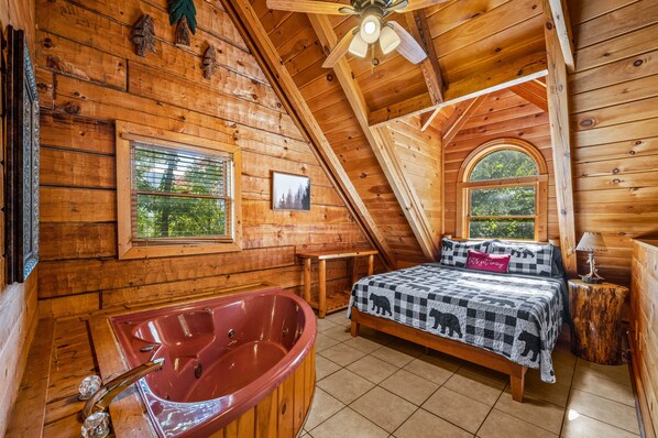 A cozy wooden cabin bedroom with a large window, a bed with a checkered blanket, a red bathtub, and a ceiling fan.