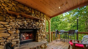 A stone fireplace with a fire, wooden deck, outdoor seating, and a view of trees. - Dog Friendly, Mtn Views, Wood Fireplaces, Hot Tub, Pool Table, Pools, Pickleball (Gatlinburg)