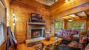 A cozy log cabin interior with a stone fireplace, wooden walls, a comfortable sofa set, and a television mounted above the fireplace. - Dog Friendly, Mtn Views, Wood Fireplaces, Hot Tub, Pool Table, Pools, Pickleball (Gatlinburg)