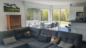 A modern living room with a grey sofa, colorful cushions, a wooden cabinet, and a large window offering a view of a patio with outdoor furniture. - Villa 4 ch - Calme- Plein Sud- SPA (Merlimont)