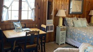 A cozy cabin interior with a wooden ceiling, a dining table with chairs, a bed with a blue floral bedspread, and a map on the wall. - Parkview Cottage #7 - 1/2 block to city beach (Ludington)