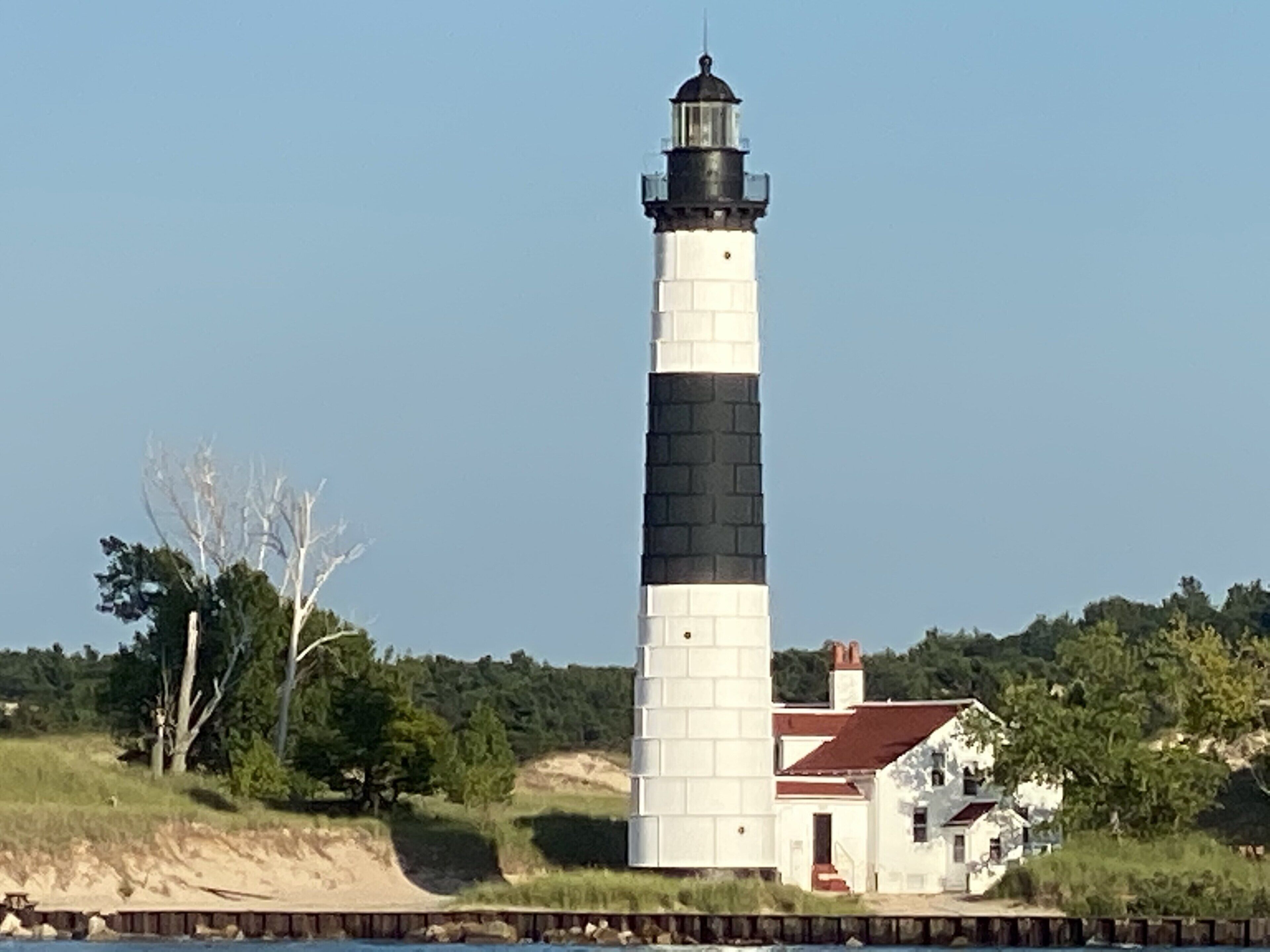 Un phare doté d’une tour rayée de noir et de blanc, un petit bâtiment adjacent et un paysage naturel composé d’arbres et d’une étendue d’eau.