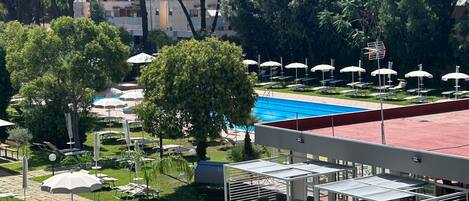 A pool area with sun loungers and umbrellas, surrounded by trees and a building with a red roof.