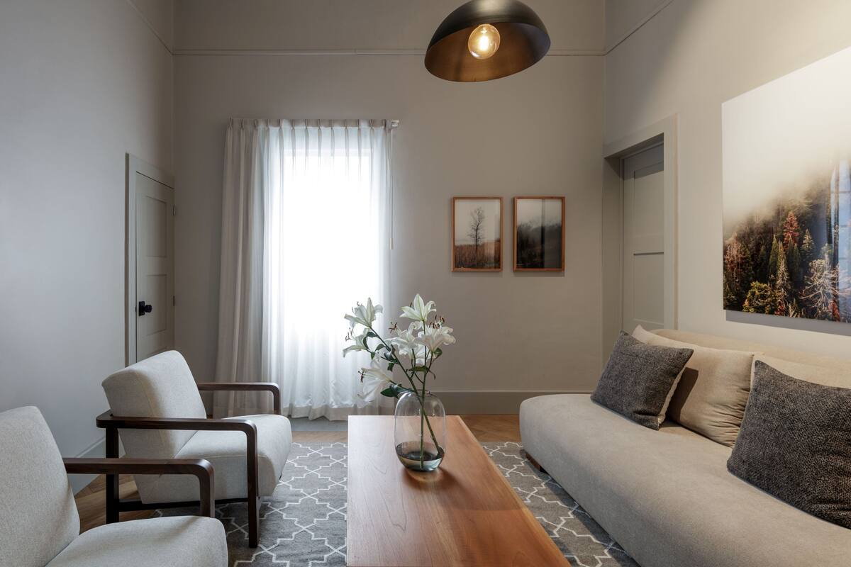 Una sala de estar moderna con un sofá, dos sillones, una mesa de centro de madera y un florero con flores. Hay dos cuadros enmarcados en la pared, una ventana con cortinas y una lámpara en el techo.