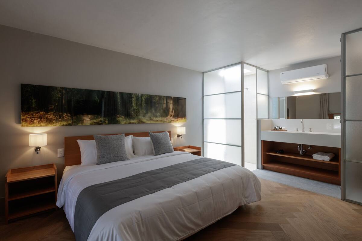 Un dormitorio moderno con una cama grande, mesitas de noche, un tocador, un ventanal amplio, un aire acondicionado instalado en la pared y un baño con lavabo y espejo.