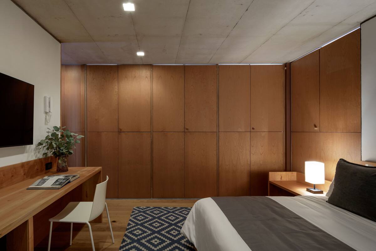 Habitación de hotel con escritorio de madera, cama, mesita de noche con lámpara y televisor montado en la pared.