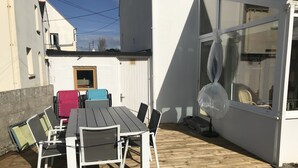A wooden deck with a table and chairs, a covered area, and a window with a chair inside. - Maison à 100m de la Plage (Treffiagat)
