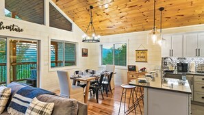 Un salón amplio con techo de madera, una mesa de comedor con sillas, y una cocina con armarios blancos y encimera negra.