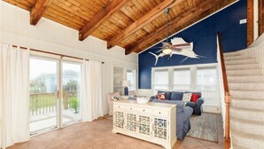 A spacious living area with a wooden ceiling, a blue accent wall, a large window, a sliding glass door, a white cabinet, and a staircase. - Oceanview Beach House - steps from the beach, really close to Galveston. TipSea! (Galveston)