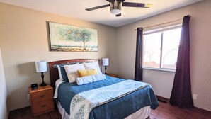 Un dormitorio con una cama, dos mesitas de noche, un ventilador de techo, una ventana con cortinas y un cuadro en la pared.
