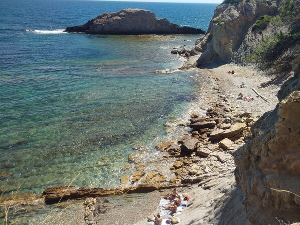 Uma praia rochosa com águas cristalinas, uma grande formação rochosa e pessoas tomando sol.