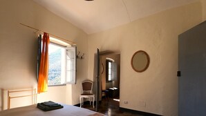 A bedroom with a bed, a chair, a window with curtains, a mirror, and a door leading to another room. - Country house near sea, ideal for families and all outdoor activities. (Finale Ligure)