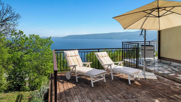 Une terrasse en bois avec deux chaises longues et un grand parasol, offrant une vue sur une étendue d’eau et des collines lointaines.