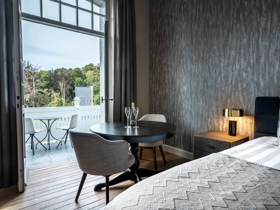 A bedroom with a bed, a bedside table with a lamp, a chair, a small table with a wine glass, and a view of a balcony with chairs and a table.