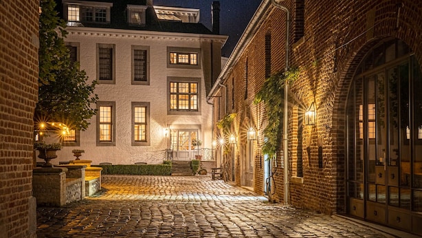 A cobblestone courtyard with brick buildings, illuminated by warm lights, and a large window on the left. - Landgoed Altenbroek (Voeren)