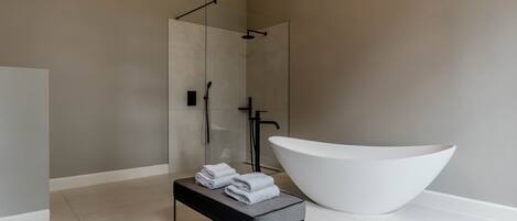 Un baño moderno con una bañera blanca grande, una ducha con cerramiento de vidrio y toallas ordenadas sobre una mesita.