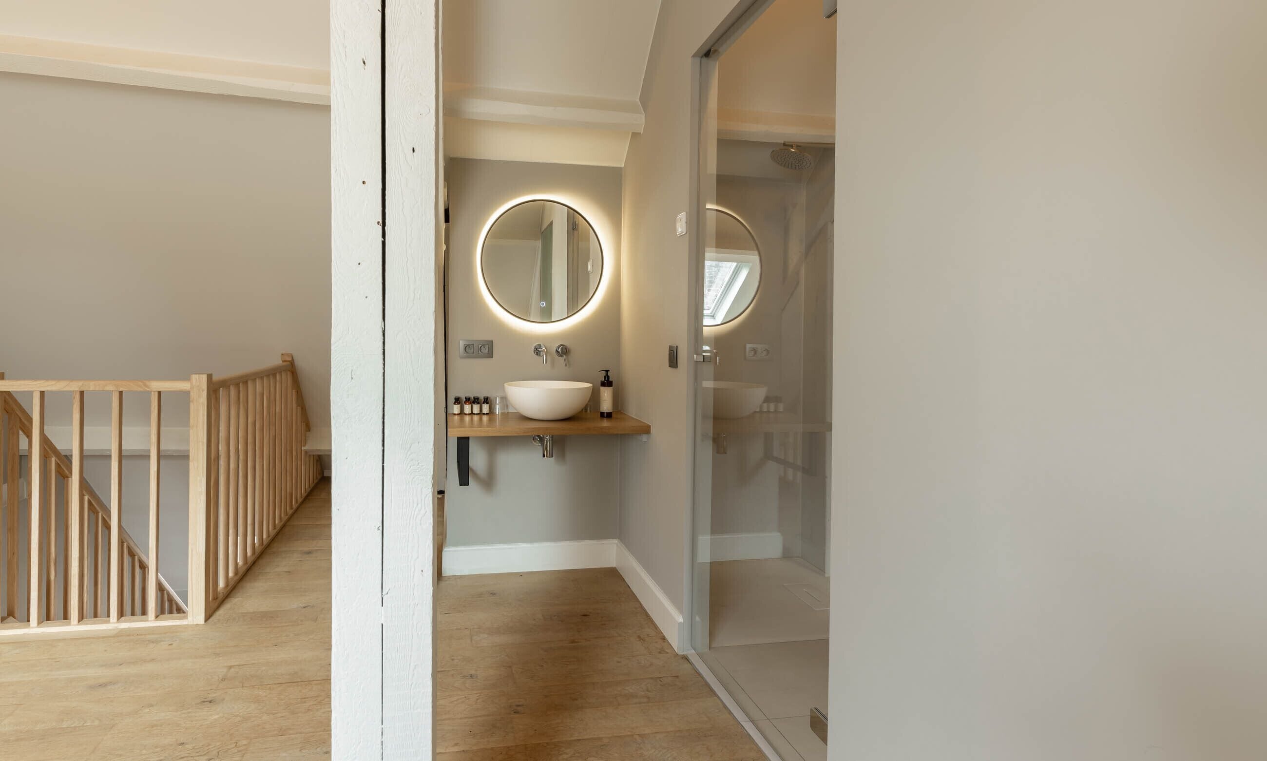 A modern bathroom with a wooden floor, a white sink with a round mirror, and a glass shower enclosure.