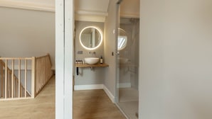 A modern bathroom with a wooden floor, a white sink with a round mirror, and a glass shower enclosure. - Landgoed Altenbroek (Voeren)