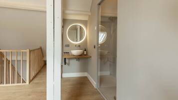 A modern bathroom with a wooden floor, a white sink with a round mirror, and a glass shower enclosure.