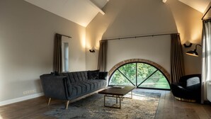 A spacious living room with a modern sofa, a coffee table, and a large arched window providing a view of greenery outside. - Landgoed Altenbroek (Voeren)