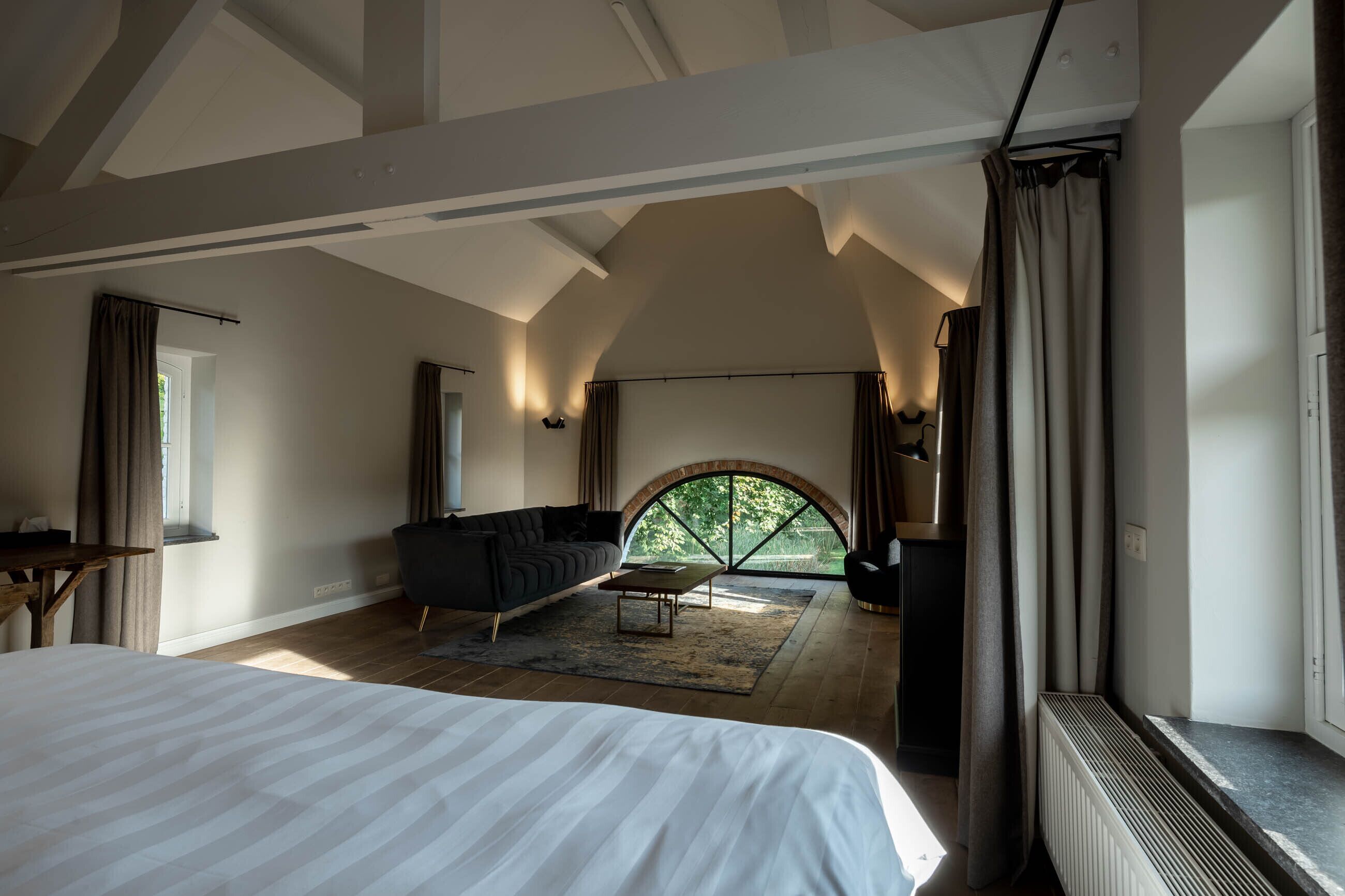Ruime slaapkamer met een groot bed, een donkere bank en een klein tafeltje. Er is een groot raam met gordijnen, en uitzicht op groen buiten.