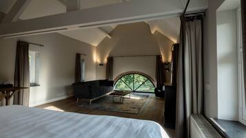 Ruime slaapkamer met een groot bed, een donkere bank en een klein tafeltje. Er is een groot raam met gordijnen, en uitzicht op groen buiten.