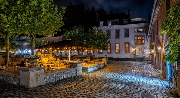 Un patio empedrado con mesas al aire libre, una fuente de piedra y un edificio con varias ventanas y un balcón.
