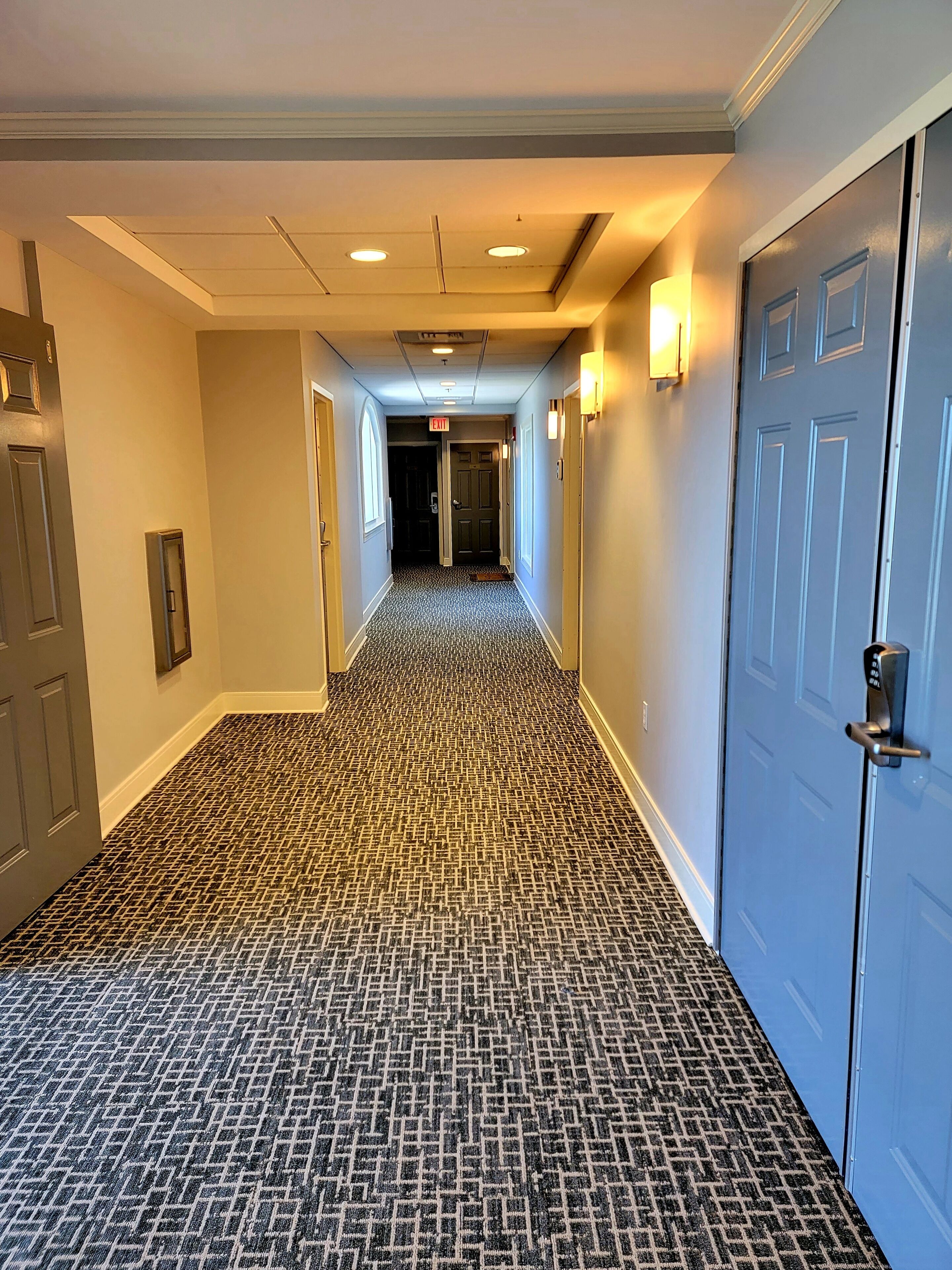 A hotel hallway with patterned carpet, doors on both sides, and recessed lighting.