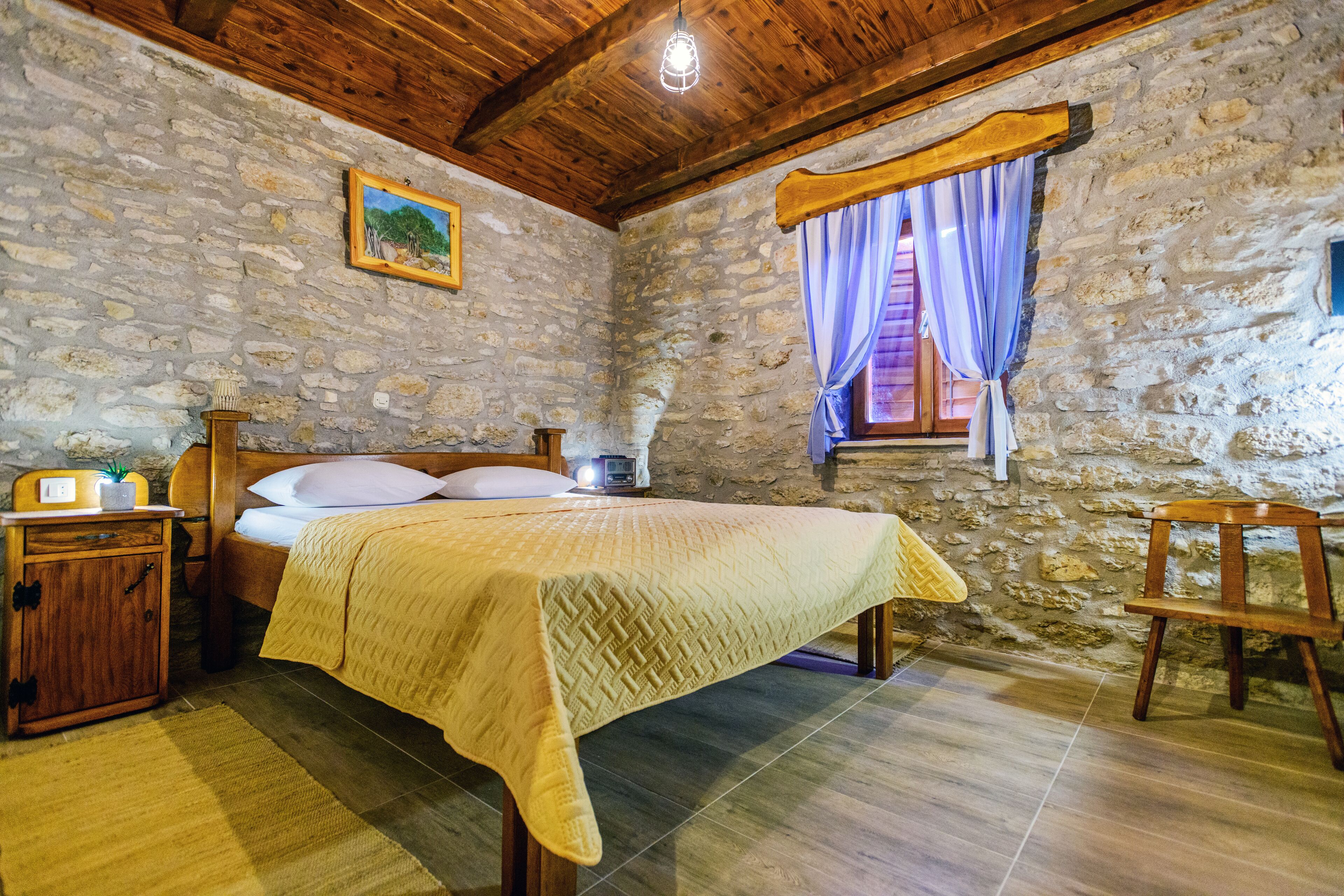 A rustic room with a stone wall, wooden ceiling beams, a bed with a yellow blanket, a nightstand with a radio, and a window with blue curtains.