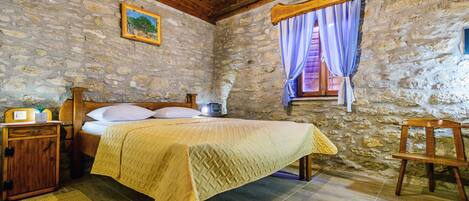 A rustic room with a stone wall, wooden ceiling beams, a bed with a yellow blanket, a nightstand with a radio, and a window with blue curtains.