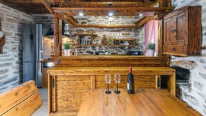 A rustic kitchen with wooden beams, stone walls, a wooden island with two wine glasses, and a bottle of wine. - Ranch Wild North (Benkovac)