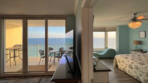 Una camera da letto con una grande finestra vista oceano, un letto con motivo a stella marina, una scrivania con televisore e un balcone con sedie e tavolo.
