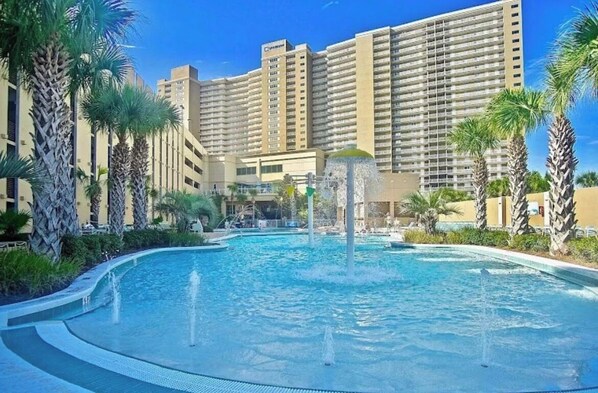 A hotel with a large pool, palm trees, and a high-rise building in the background.