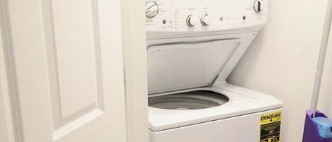 A laundry room with a front-loading washer and dryer, a door, and a purple cleaning tool on a shelf.