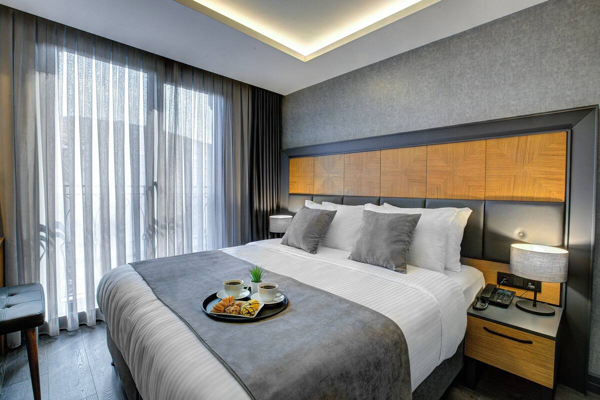 A modern hotel room with a large bed, bedside table with a lamp, and a tray with breakfast items.