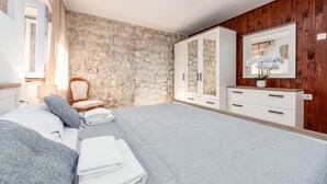 Un dormitorio con pared de piedra, una cama con ropa de cama gris, un armario de madera y un espejo con una orquídea en maceta.