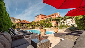 Un área junto a la piscina con sillones de descanso, una mesa de vidrio con bebidas y una sombrilla roja.