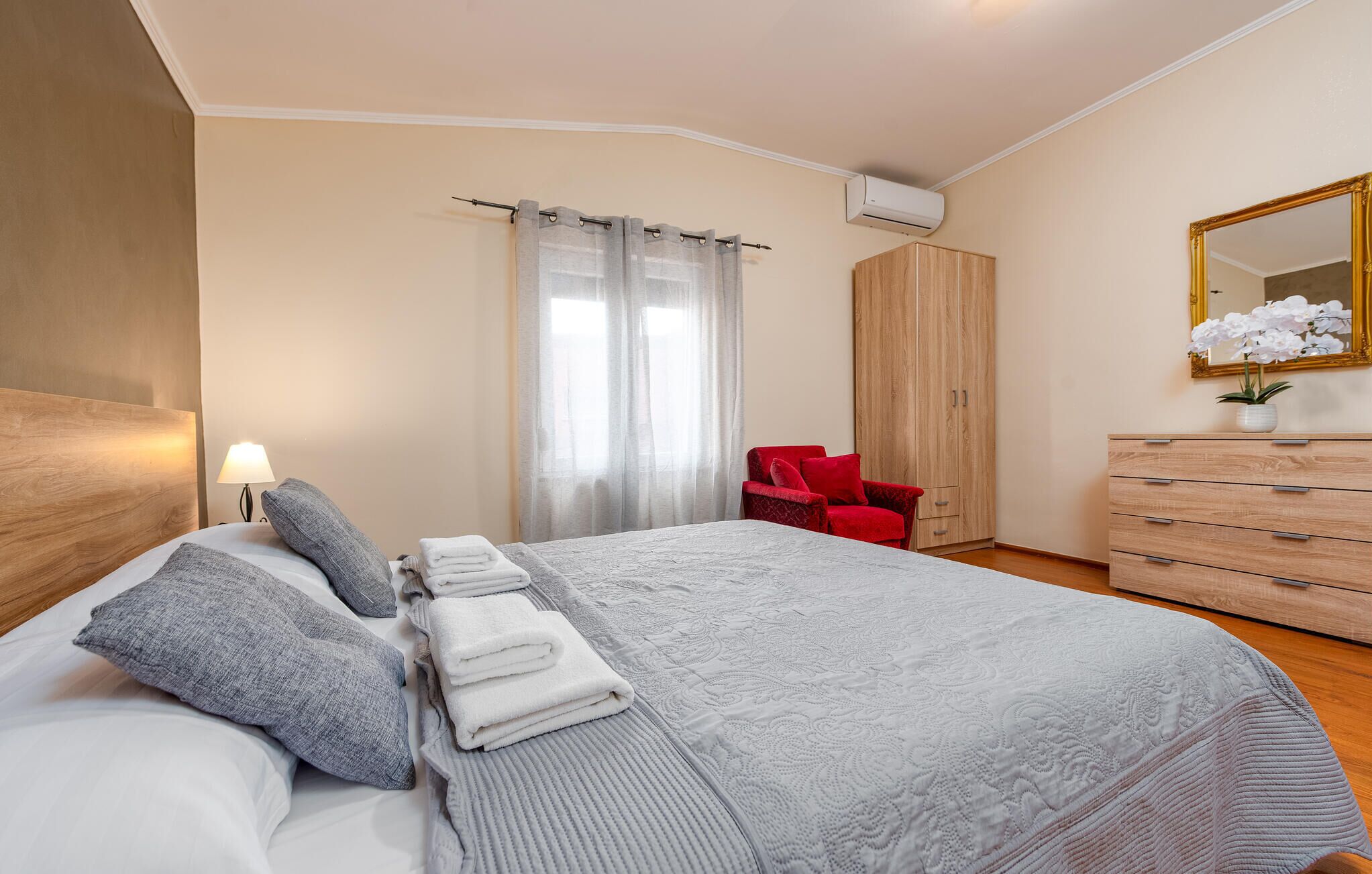 A hotel room with a bed, a red chair, a wooden wardrobe, a dresser, and a mirror.