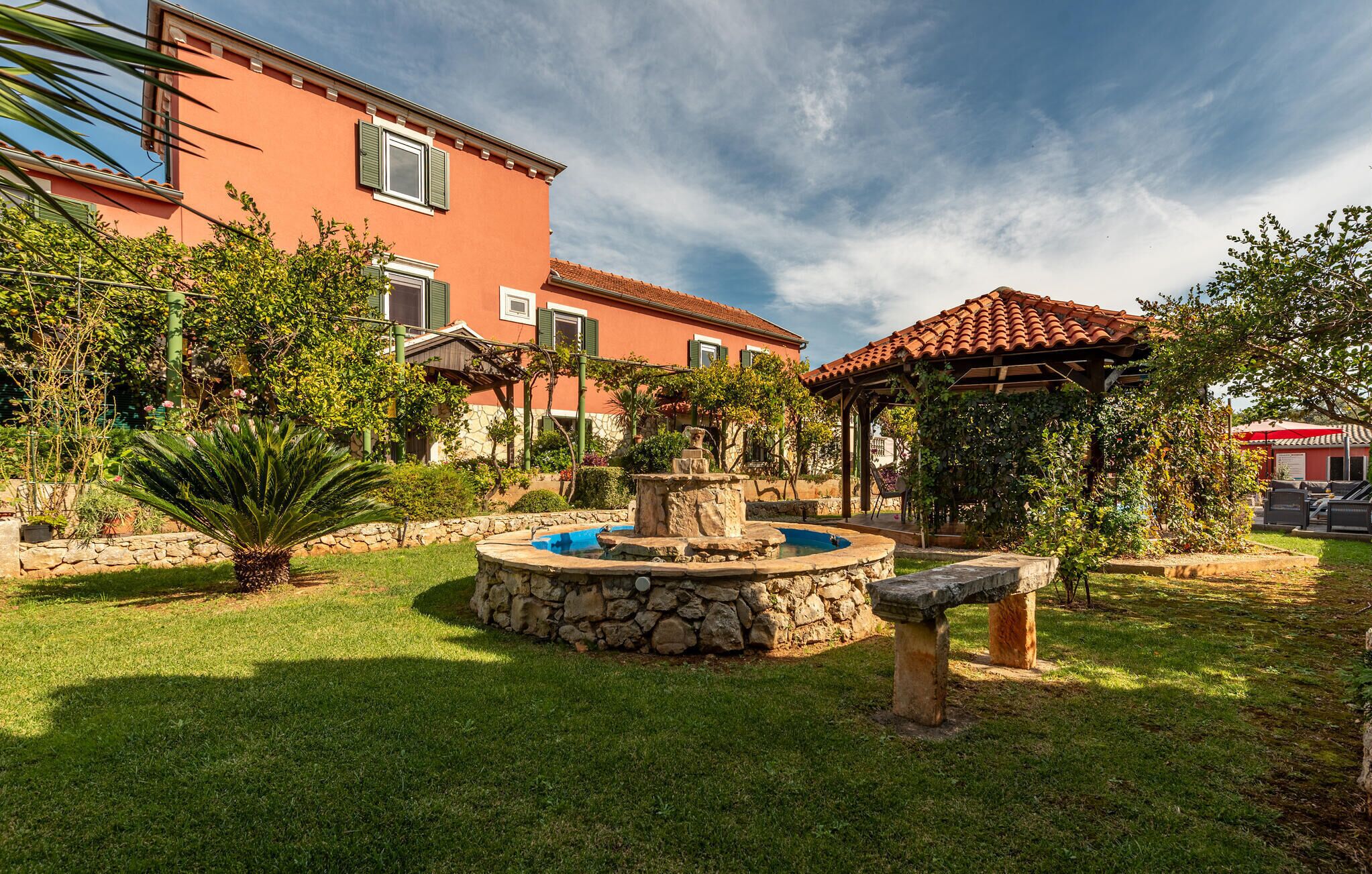A courtyard with a stone fountain, lush greenery, and a building with a red facade and green shutters.