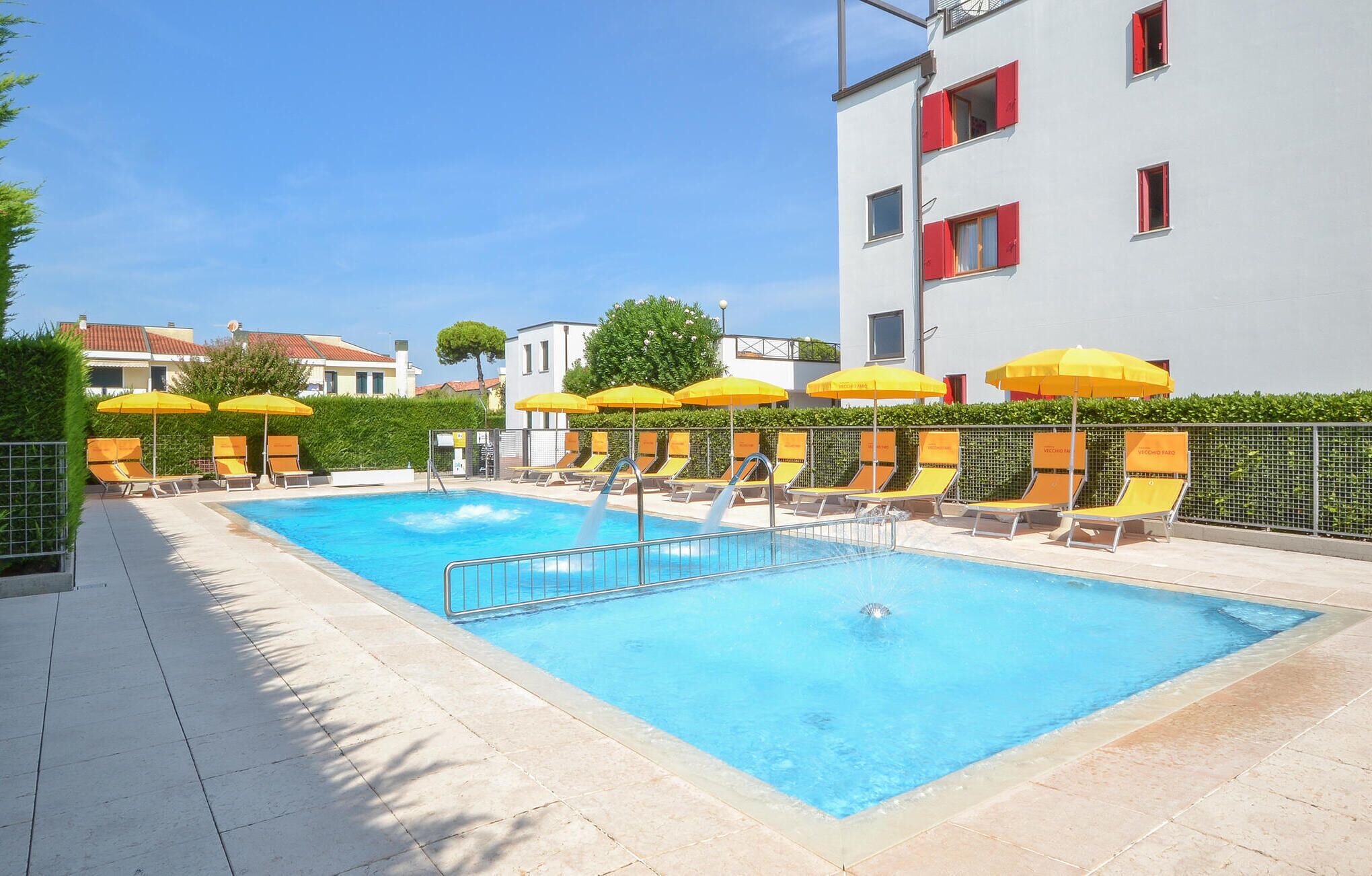 A swimming pool with sun loungers and yellow umbrellas, surrounded by a fence and a building with red shutters.